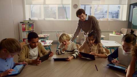Sáu em học sinh cầm máy tính bảng ngồi tại bàn học chung và một giáo viên xem quá trình hoạt động của một em học sinh